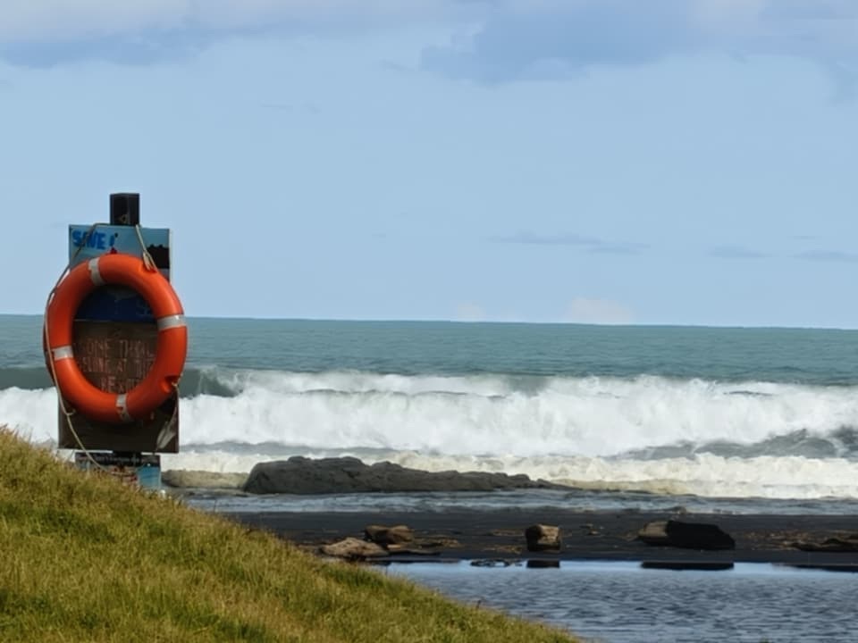 An orange lifebuoy on its stand above a black sand beach as surf rolls in